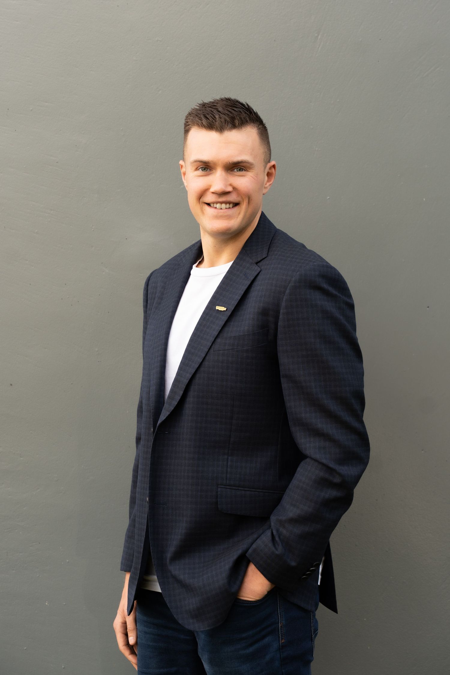Smiling young man in navy blazer and white t-shirt posing confidently against a gray wall.