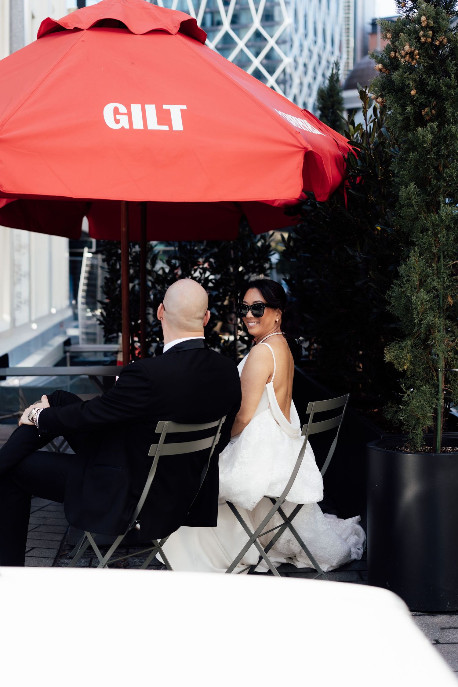 Bride and groom sitting at outdoor cafe table under red Gilt umbrella in wedding attire, urban setting.