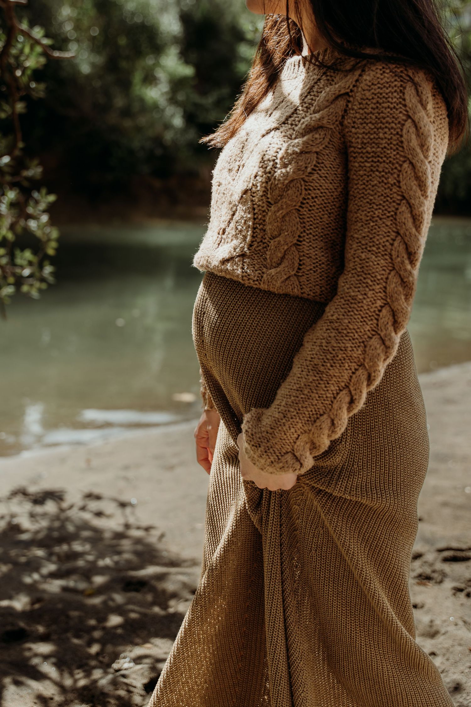 Pregnant woman in beige cable-knit sweater and ribbed skirt standing near a serene river.
