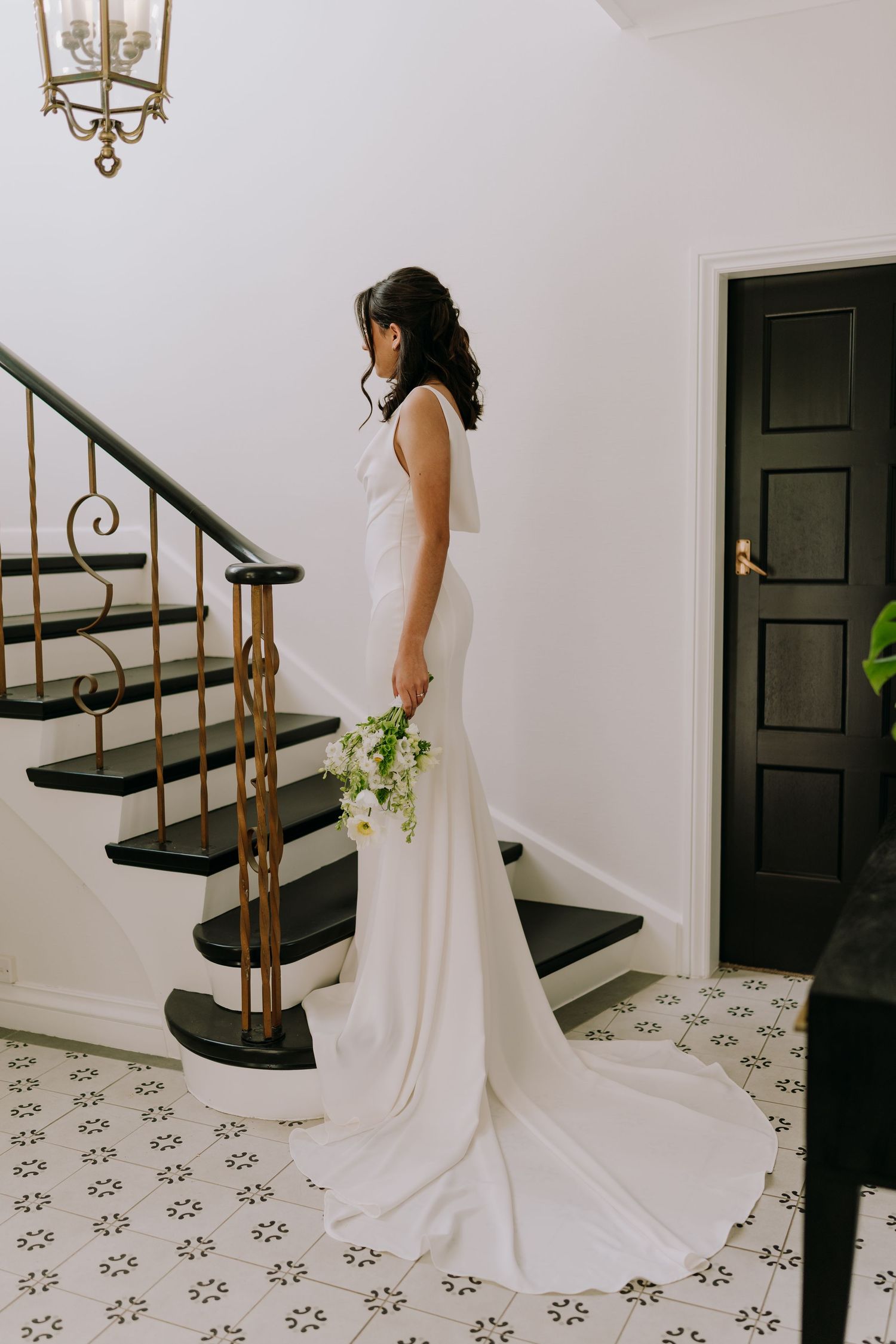 Bride in sleek white wedding gown stands on patterned tile floor near black door and ornate staircase.
