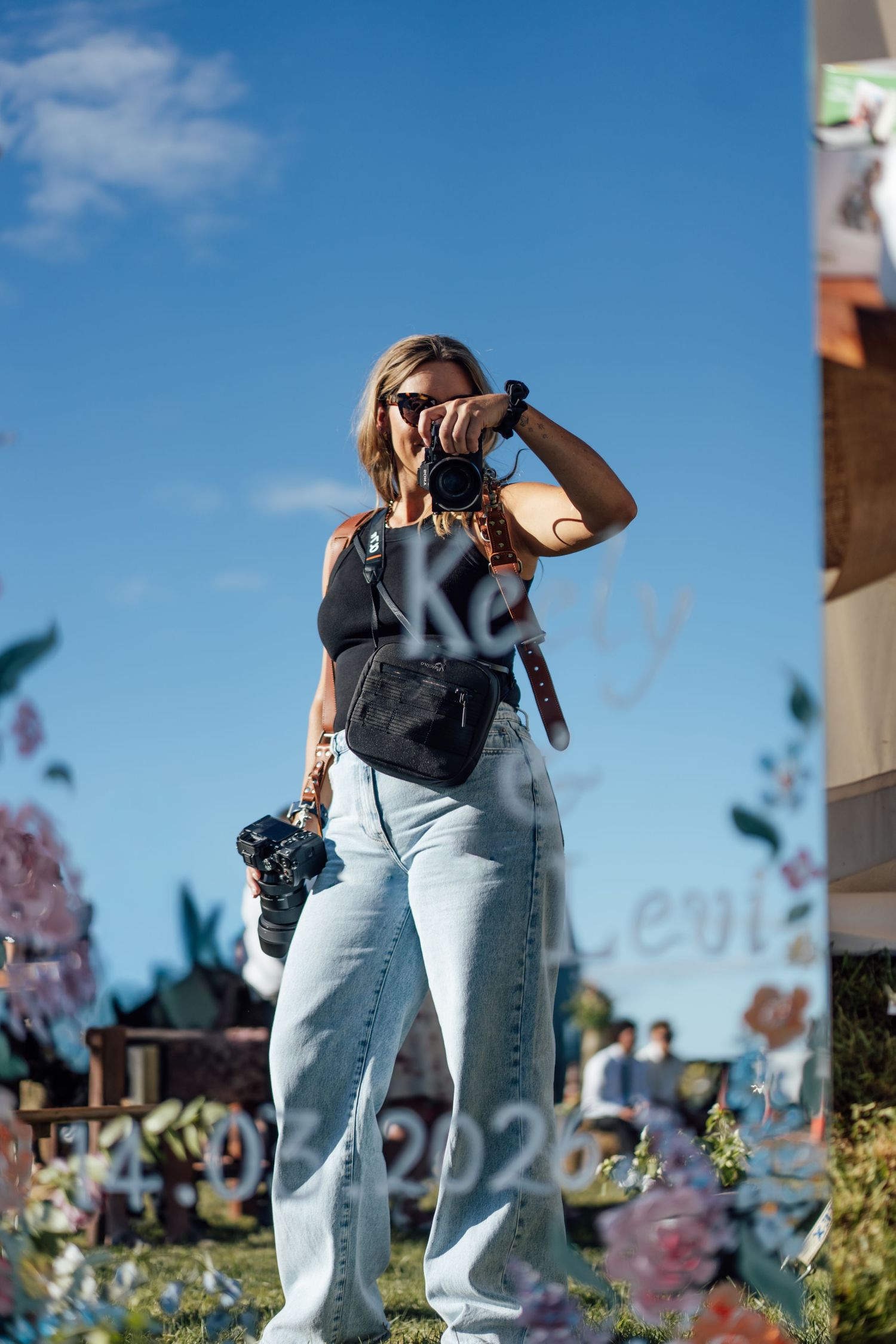 Woman photographer in jeans and black top holding camera outdoors at a floral event under blue sky.