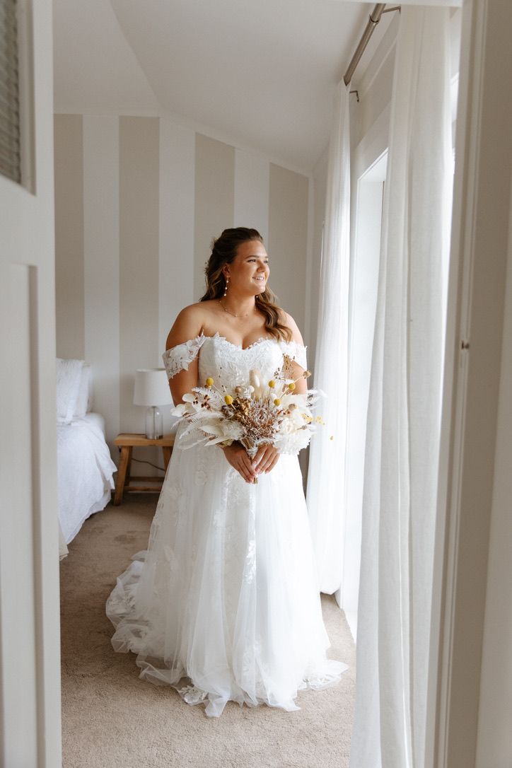 Bride in flowing off-shoulder wedding dress walks through bright white hallway with natural light.