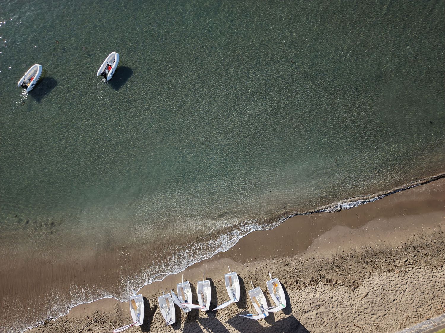 Aerial view of small boats moored near a sandy beach with clear turquoise water and sailboats lined on shore.