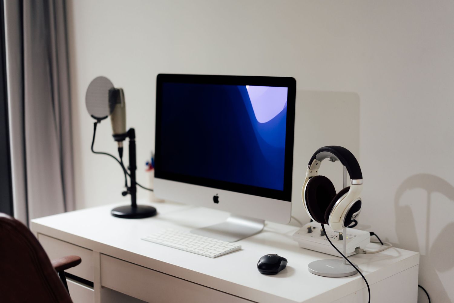 White desk setup with iMac, USB microphone, wireless headphones on stand, and Apple keyboard and mouse.