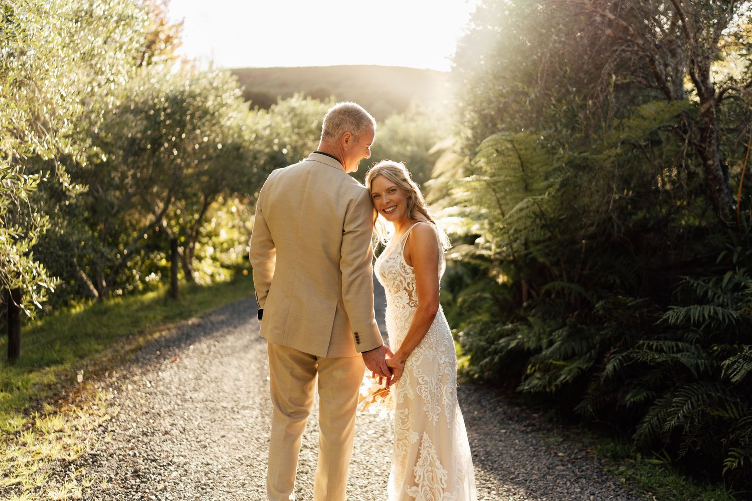 Bride and groom holding hands on a sunlit gravel path surrounded by lush greenery on their wedding day.