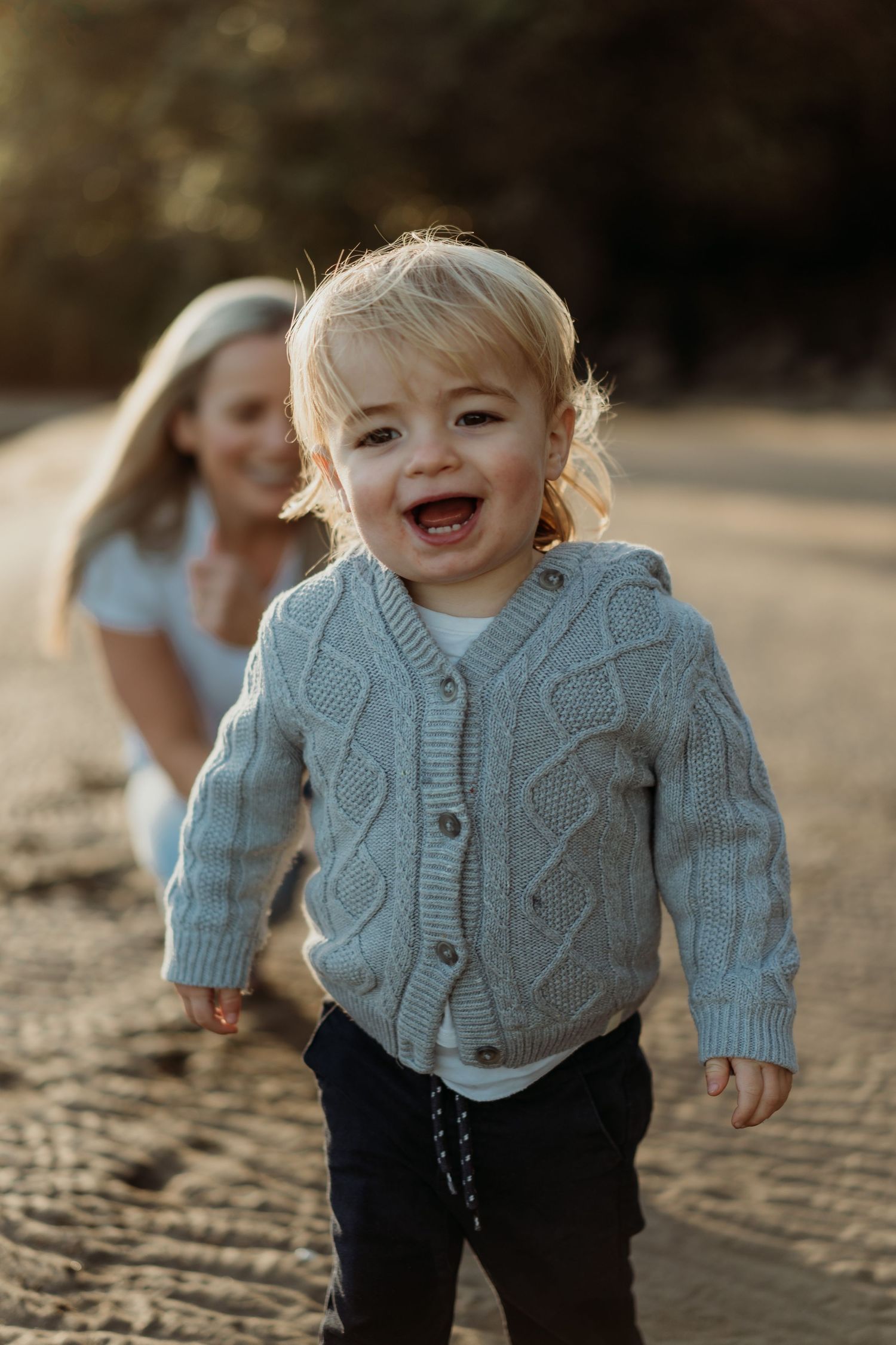 Happy toddler in gray knit cardigan laughing outdoors with smiling mother in soft golden light background.