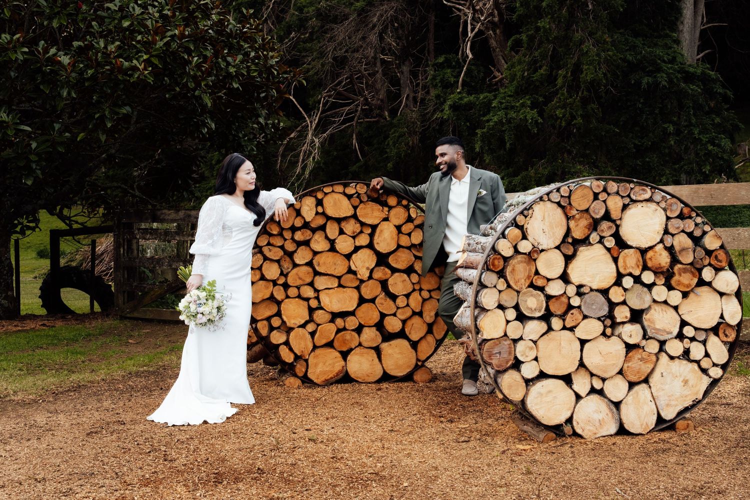 Bride and groom posing beside large circular stacked log piles outdoors on their wedding day.