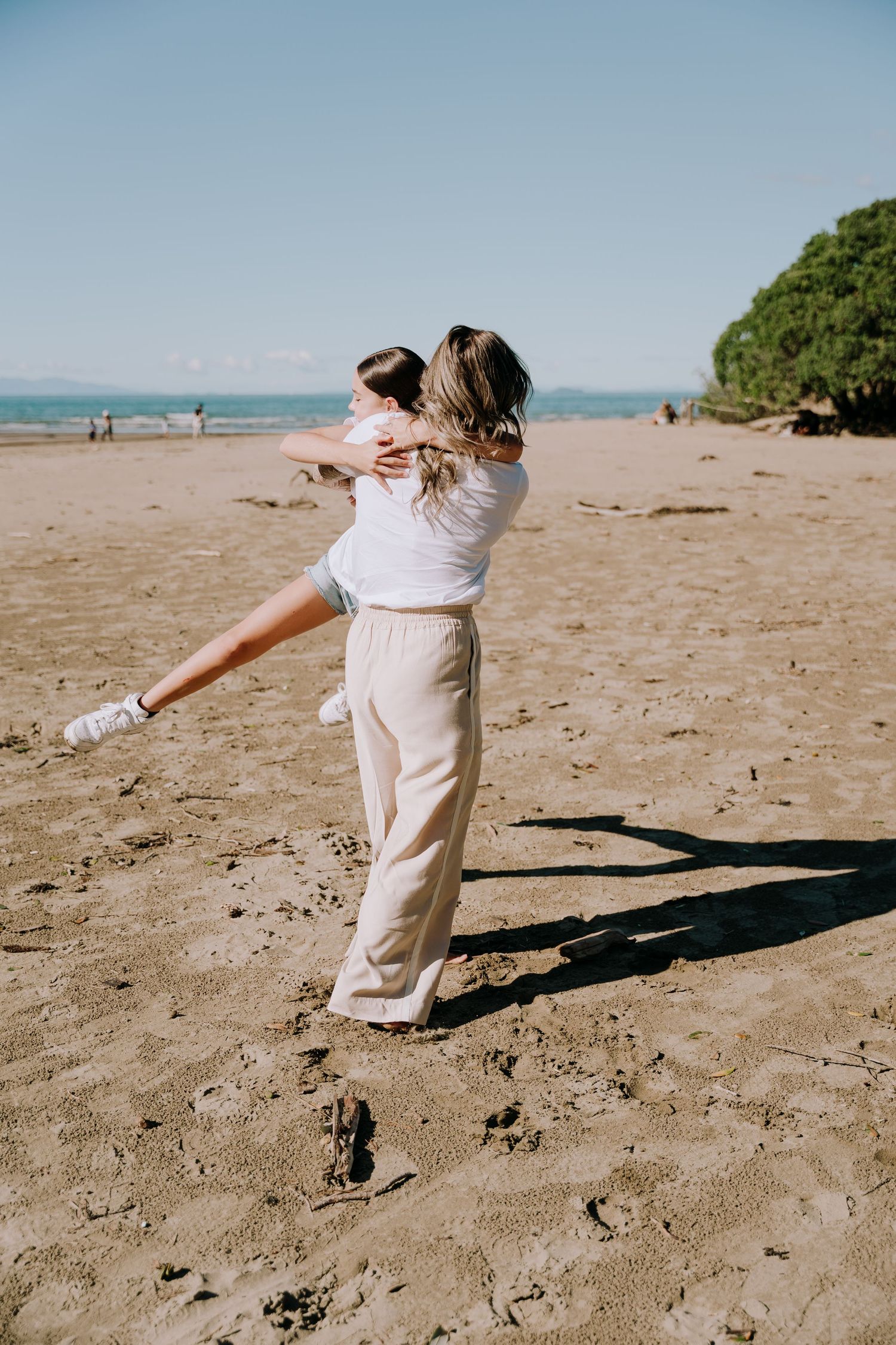 Person in white top and beige pants spins joyfully on a sunny beach with ocean and trees in the background.