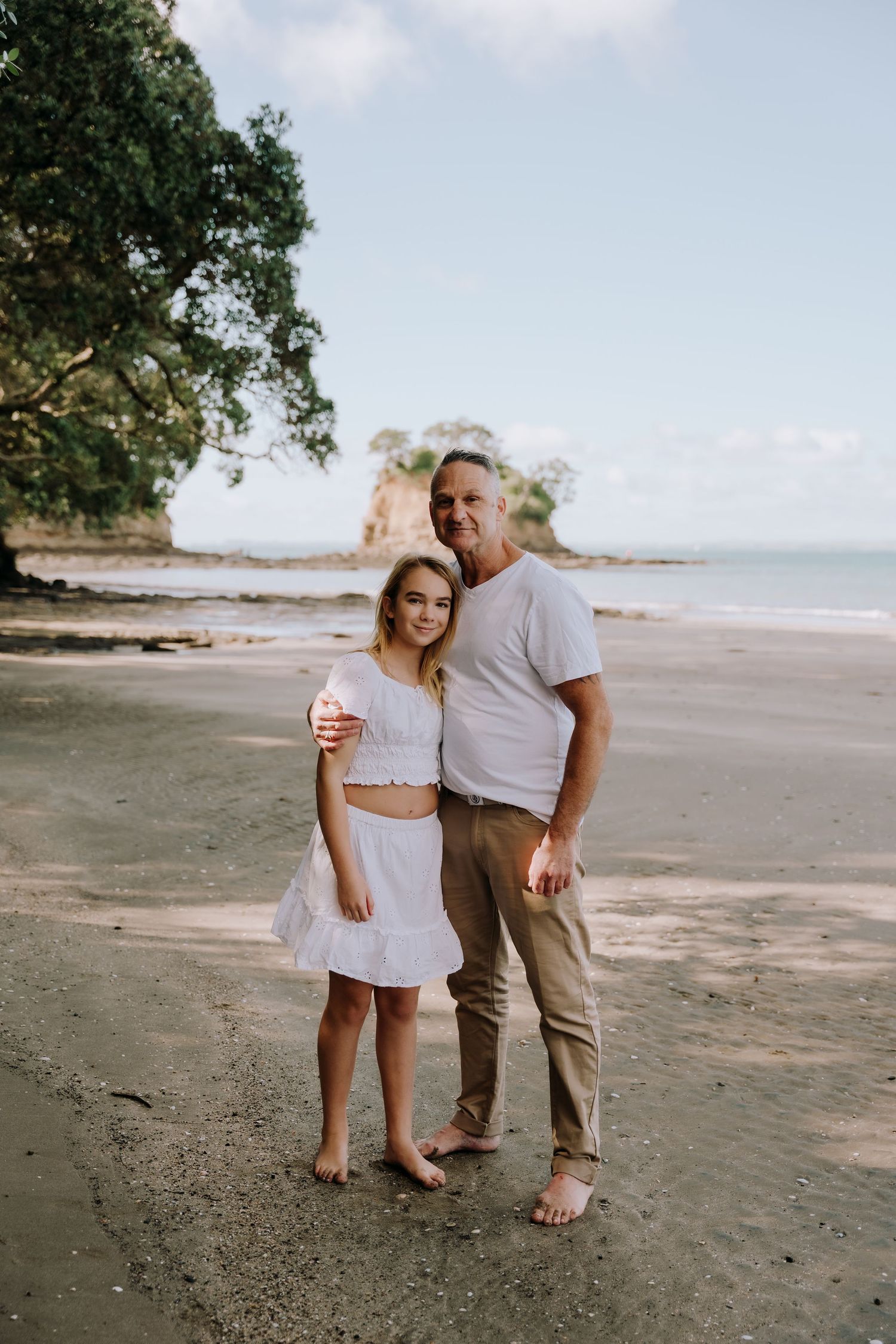A couple shares an intimate moment while walking along a scenic beach with trees in the background.