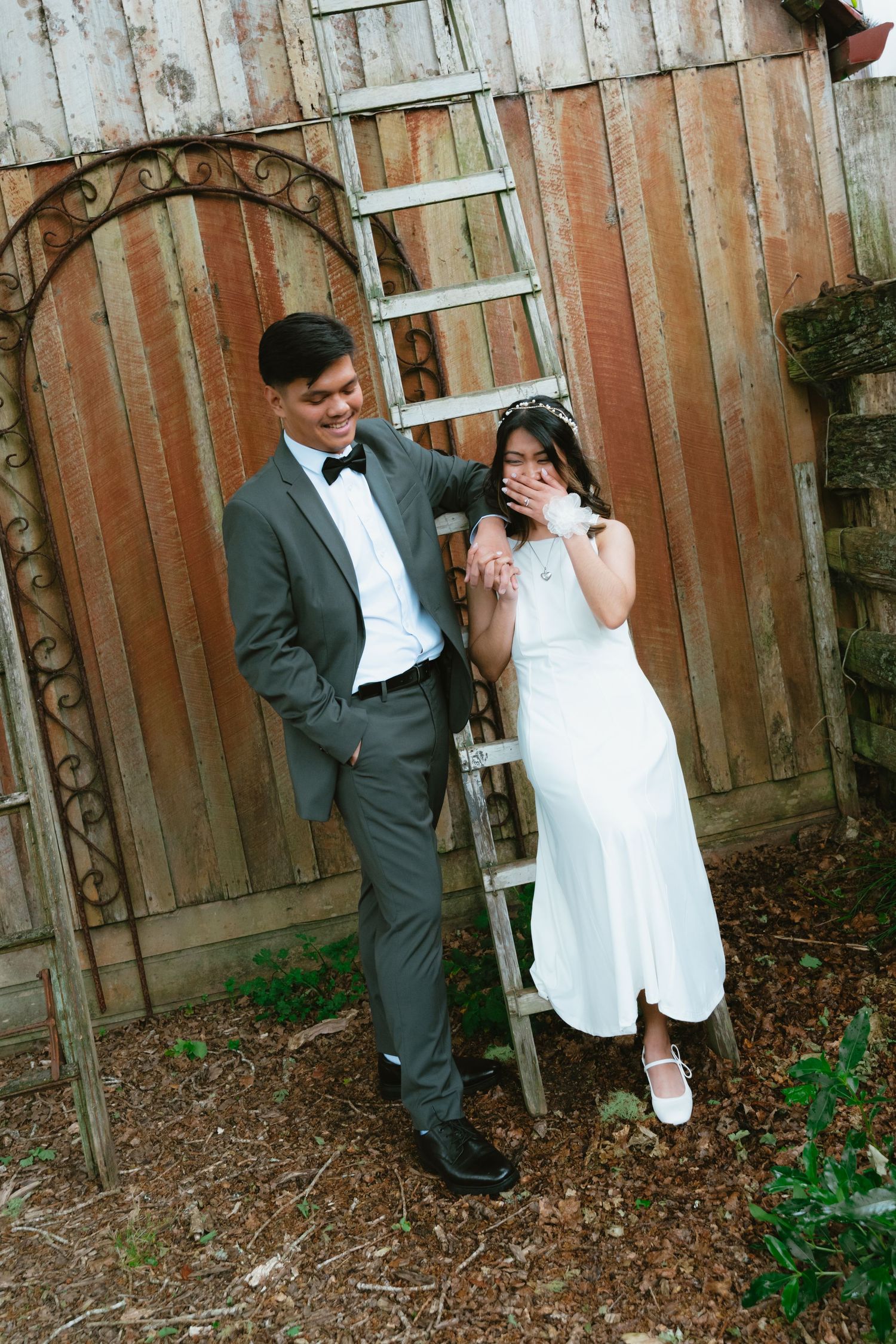 Playful wedding couple poses against rustic wooden fence in a garden setting.