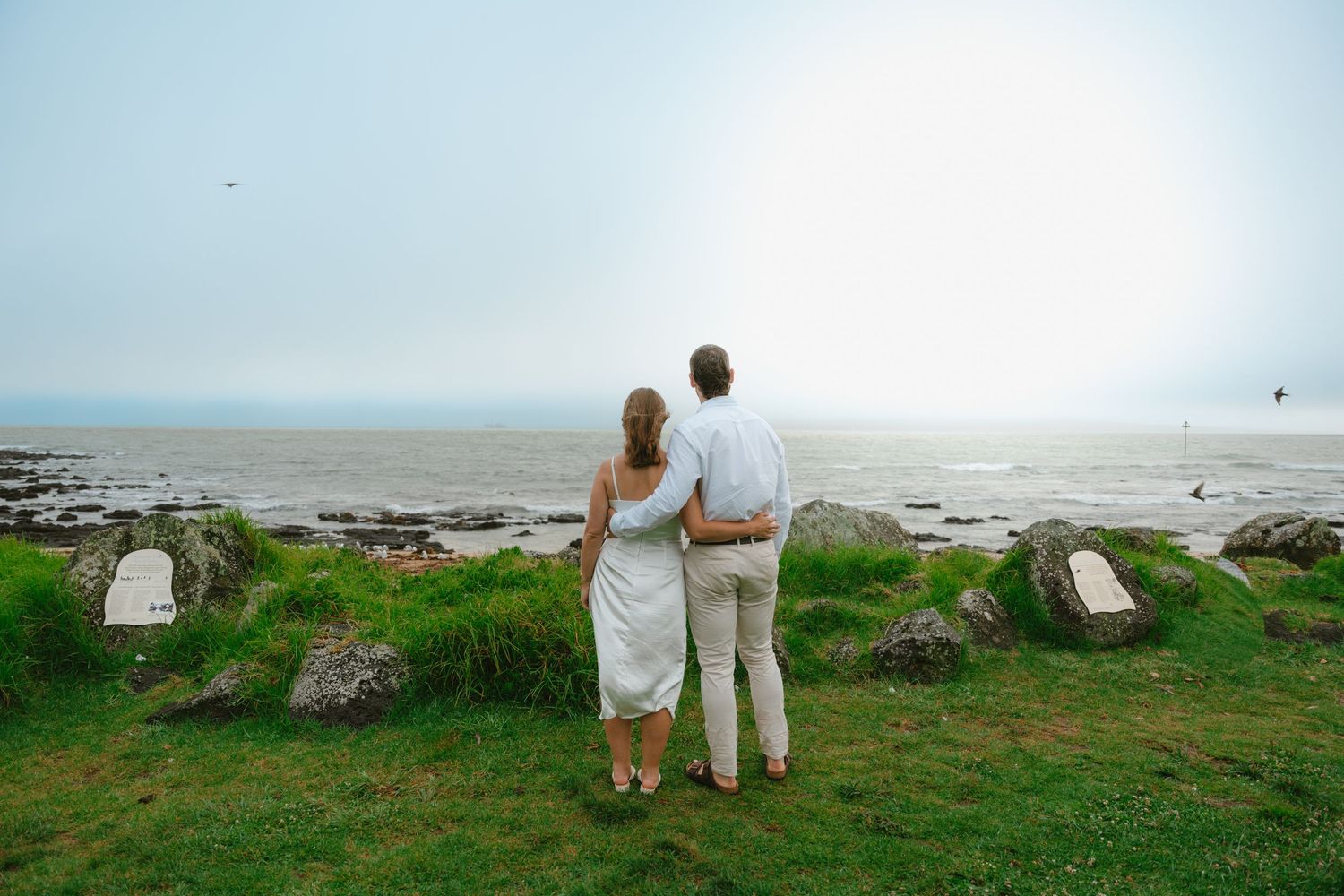 Two people in white clothing stand together looking out over a scenic coastal landscape at sunset.