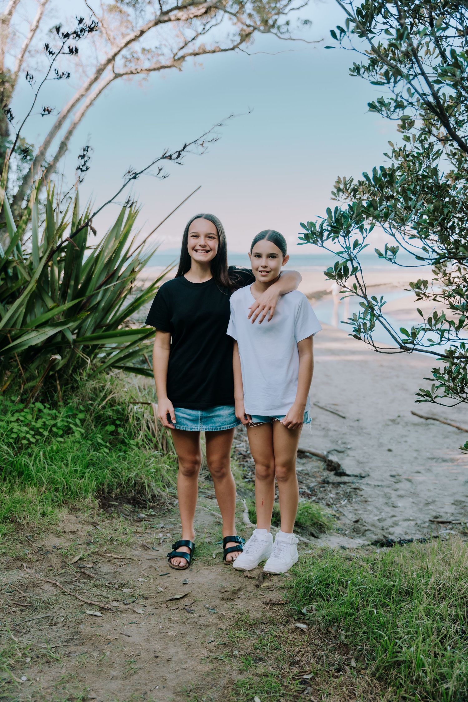 Two friends stand together on a coastal path with tropical plants and ocean views at sunset.