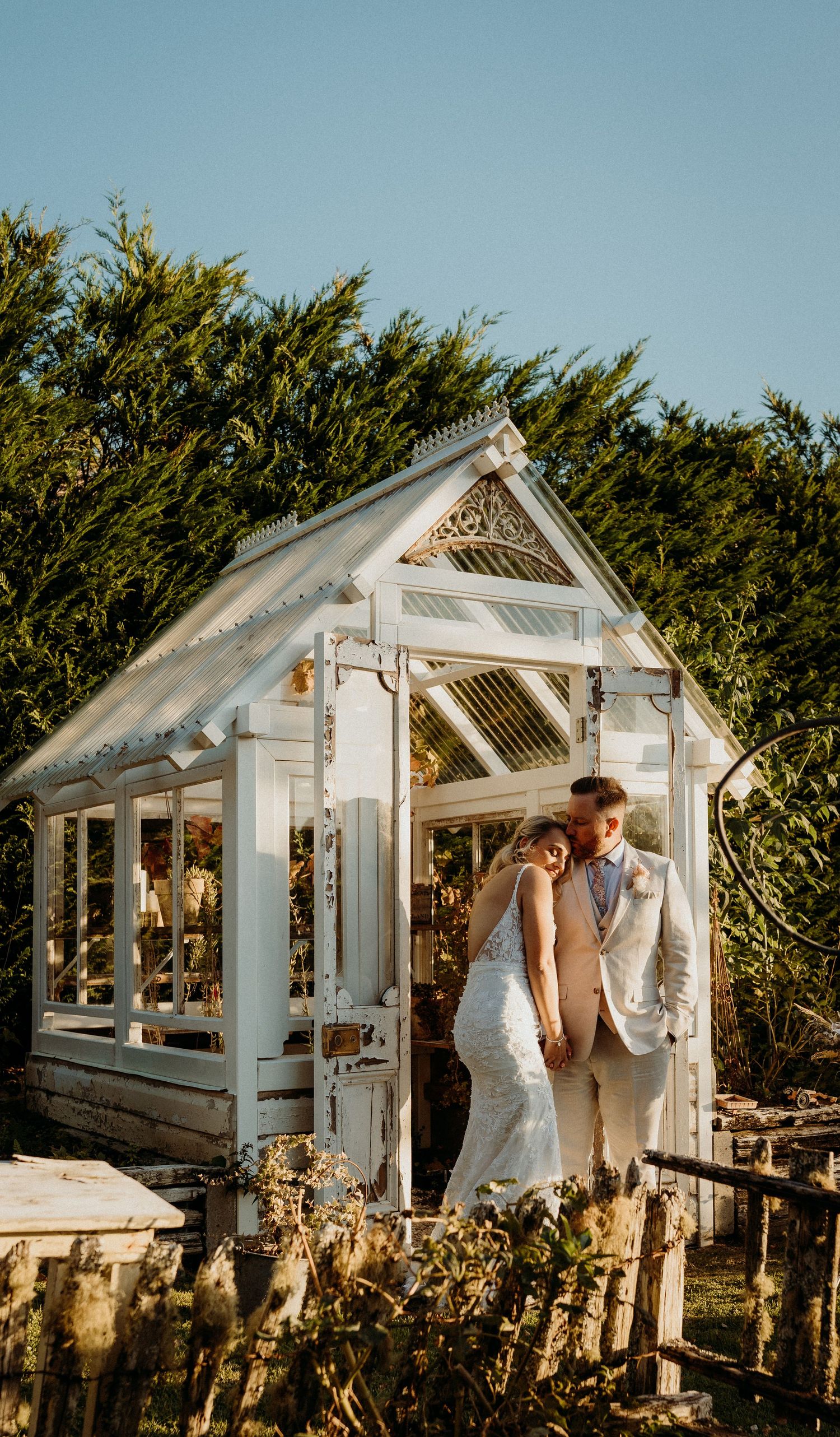 Couple shares intimate moment in white decorative greenhouse surrounded by lush greenery on sunny day.