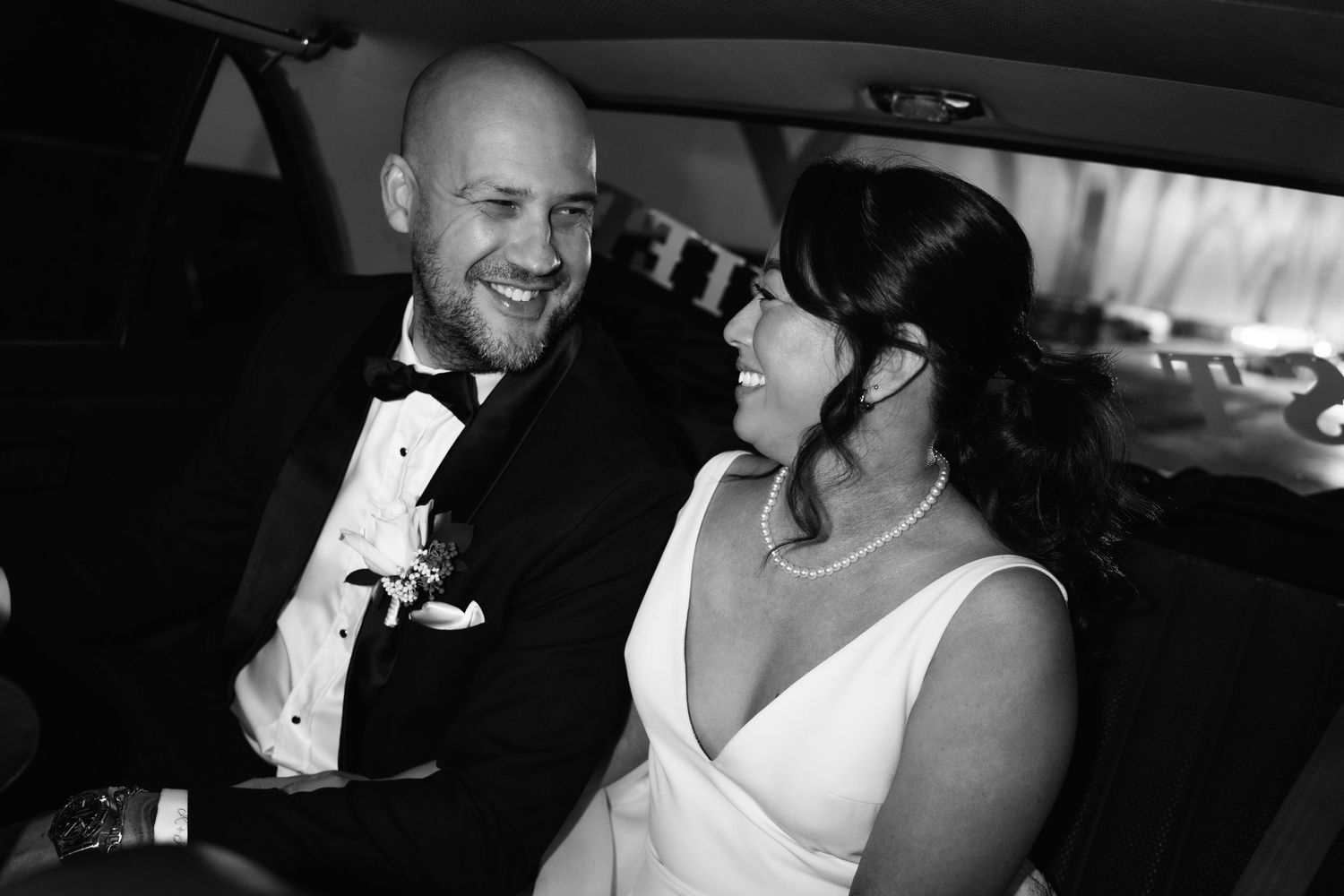Black and white photo of a smiling bride and groom in formal wedding attire sitting together in a car.
