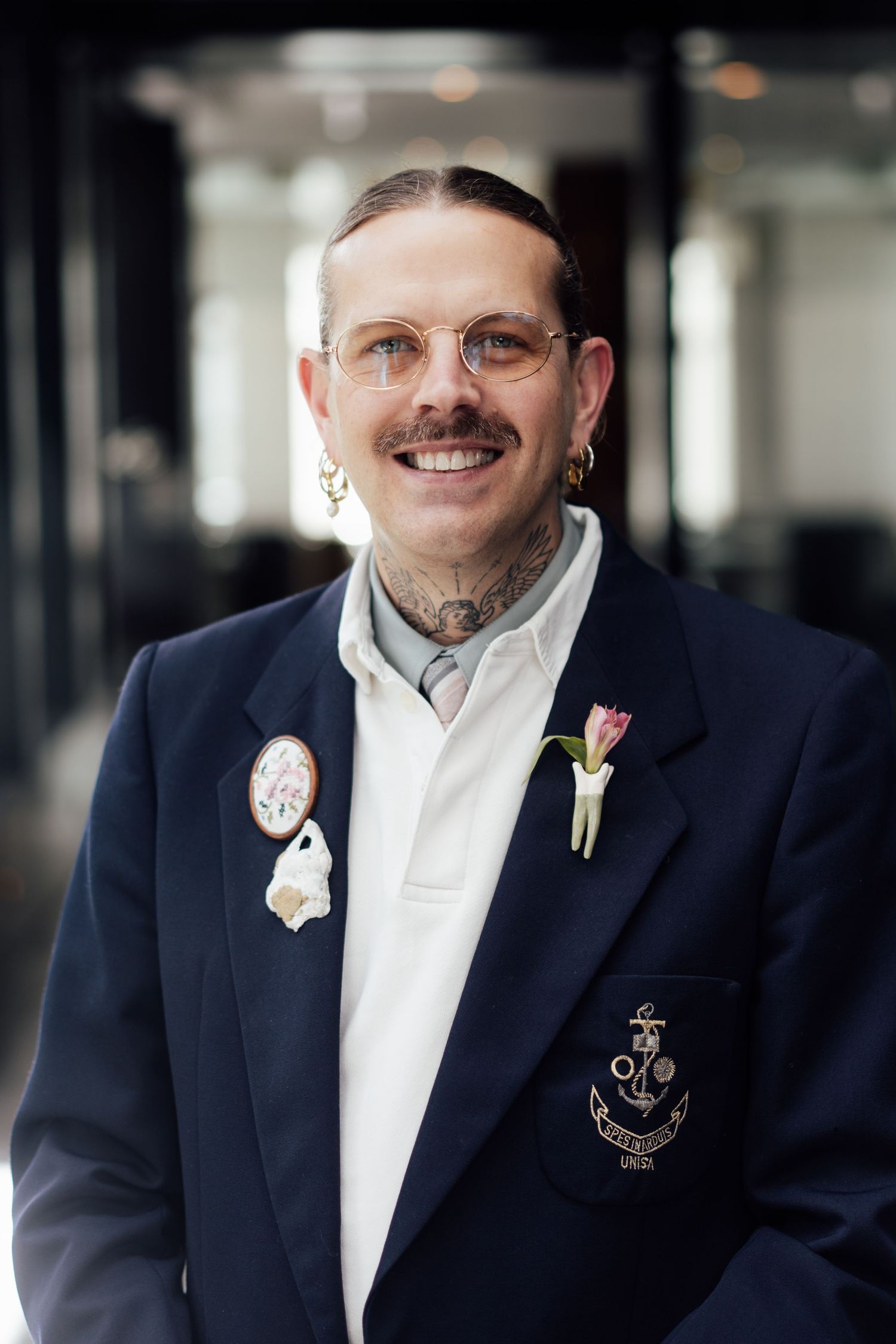Smiling man with tattoos and mustache wearing a navy blazer with pins and a floral buttonhole.