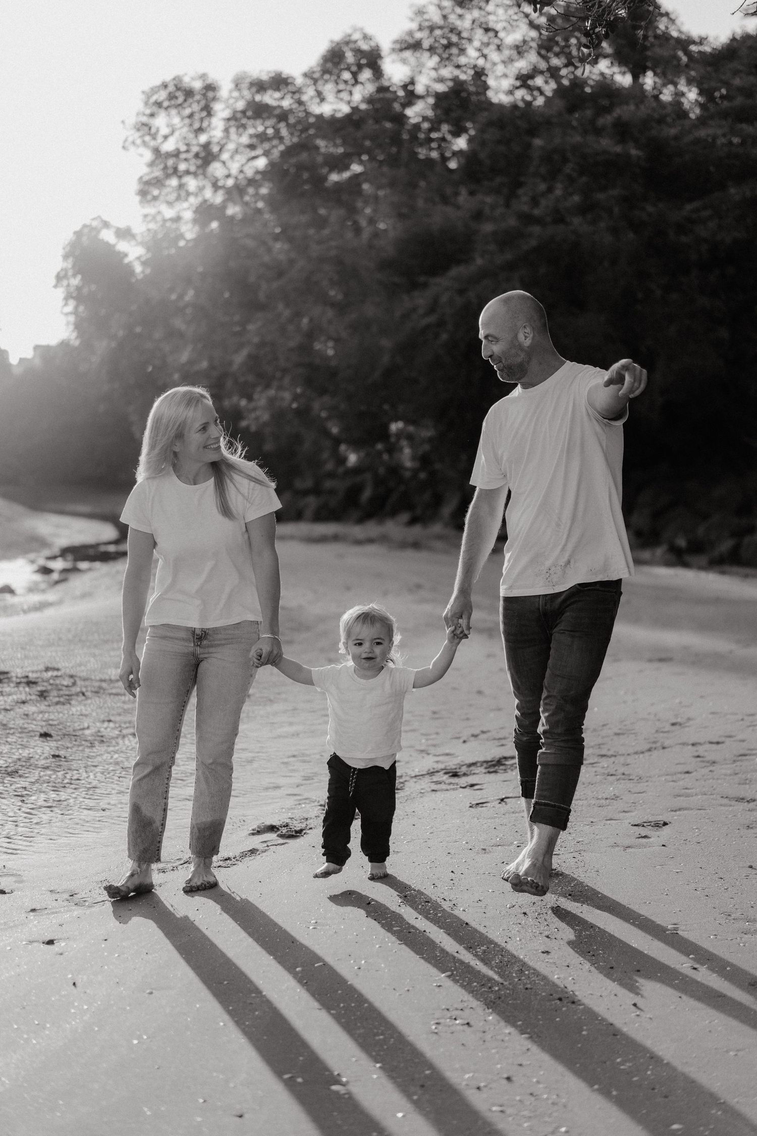 Black and white photo of a family with a toddler walking on a beach, parents holding child's hands.