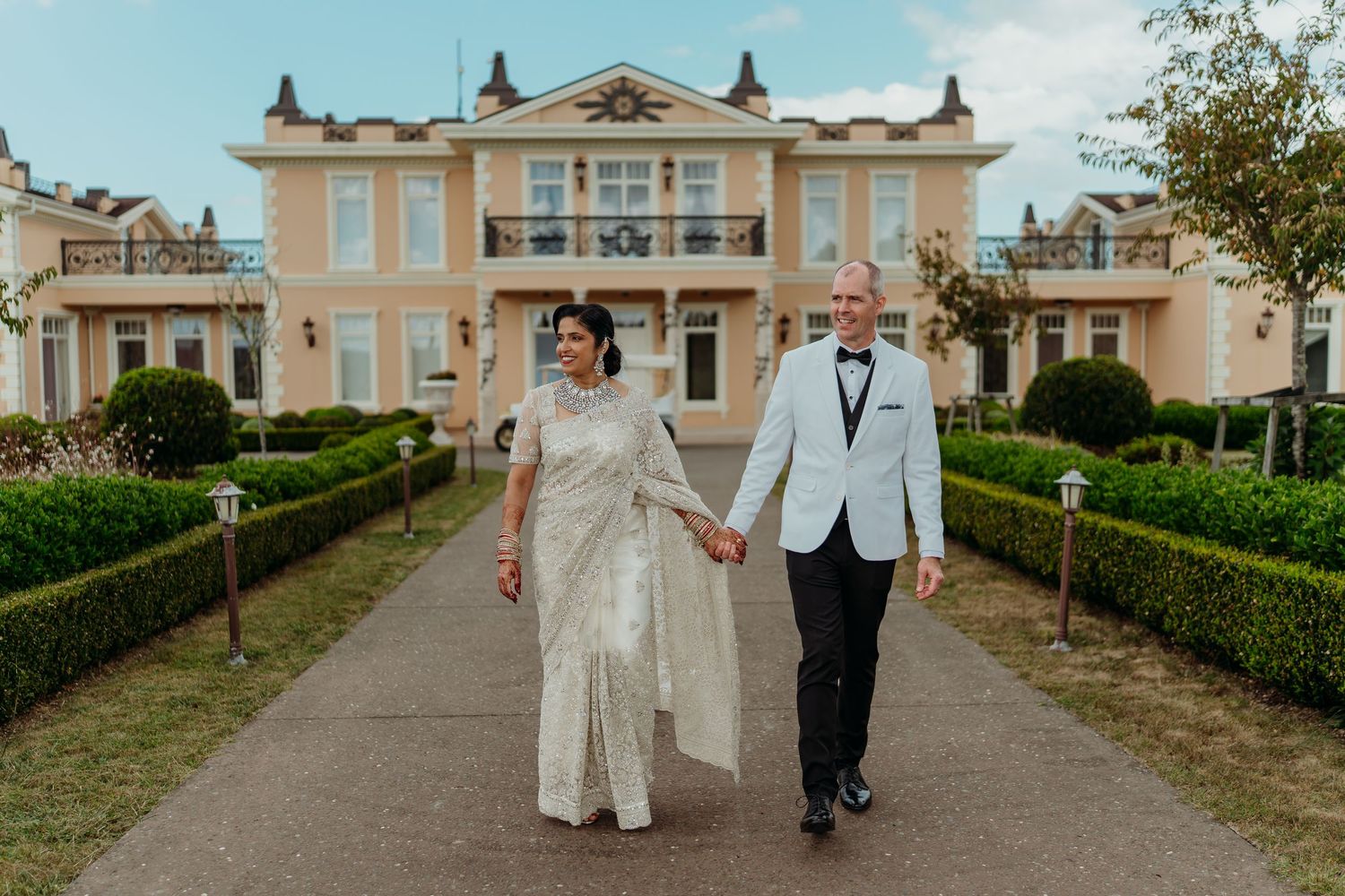 Couple in elegant wedding attire holding hands walking toward a grand yellow mansion with manicured hedges.