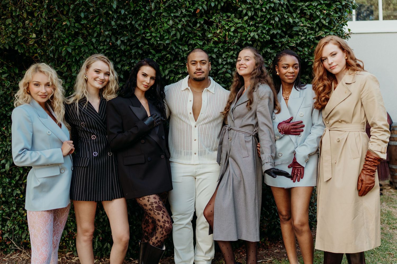 Seven stylish models pose together outdoors in elegant fashion outfits, showcasing diverse styles from blazers to trench coats.