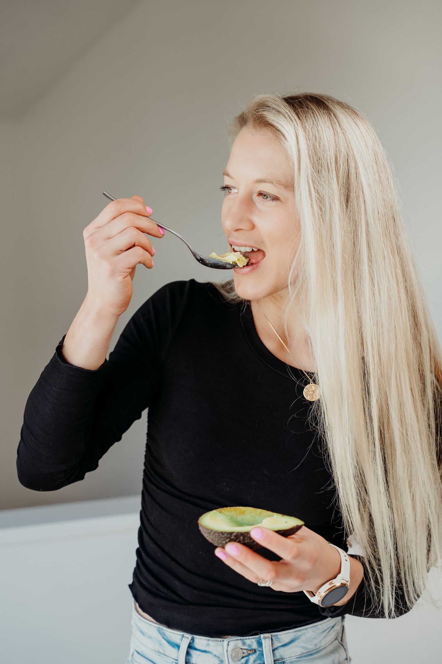 Smiling blonde woman in black top eating avocado with a spoon, holding halved avocado, healthy eating concept.