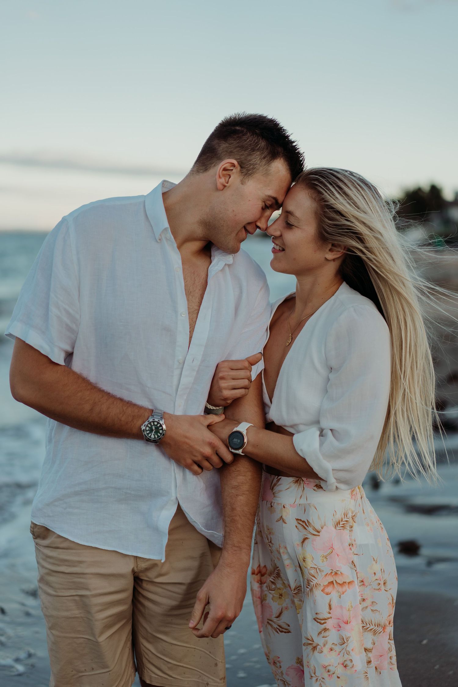 Romantic couple in white clothing share an intimate moment on the beach at sunset with wind blowing through her blonde hair.