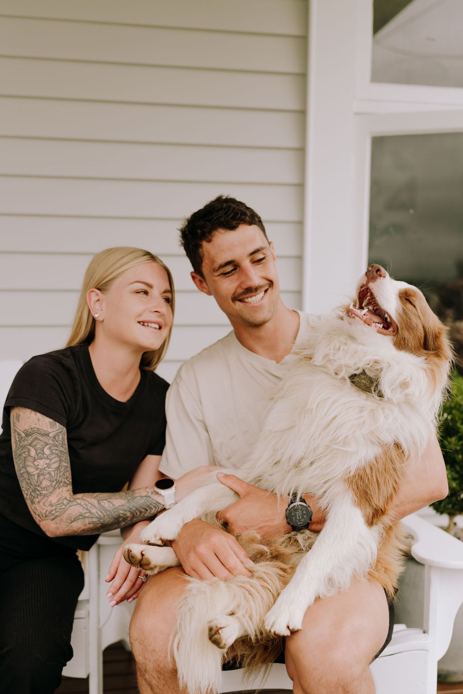 A happy couple laughing with their playful dog on a white porch chair.