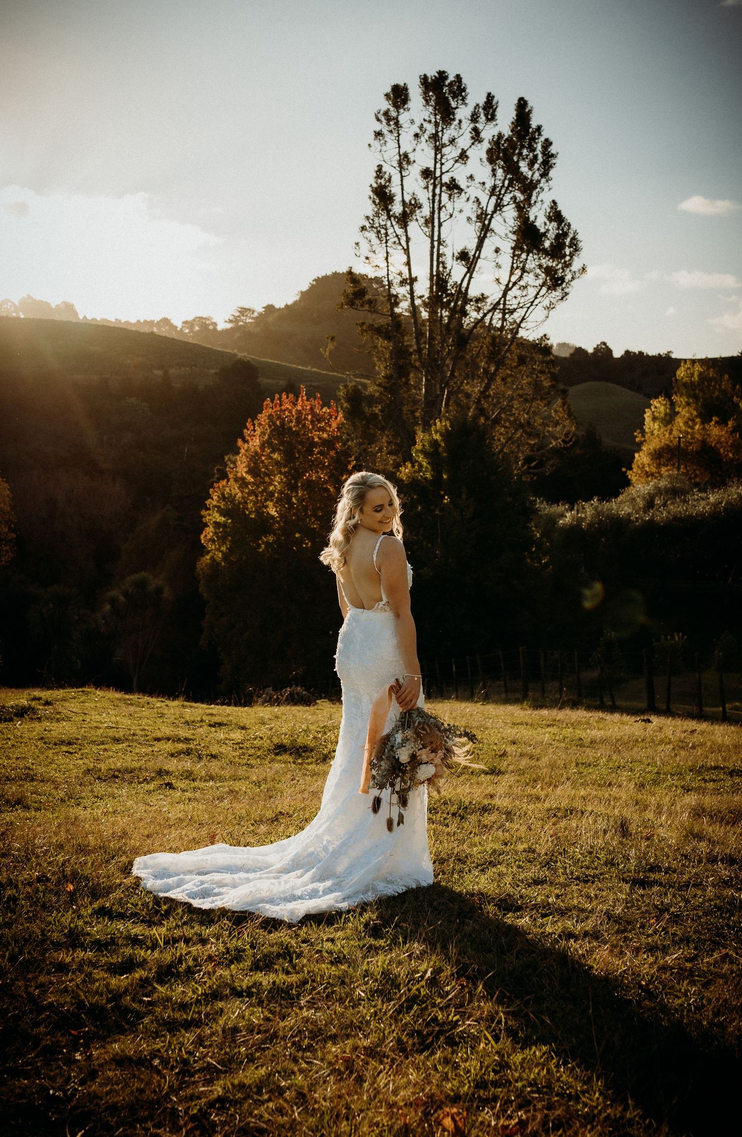 Person in white wedding dress stands in golden sunlight holding bouquet against autumn landscape.