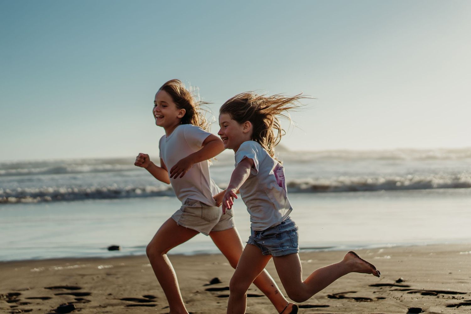 Two young girls running joyfully along a sandy beach with ocean waves in the background on a sunny day.