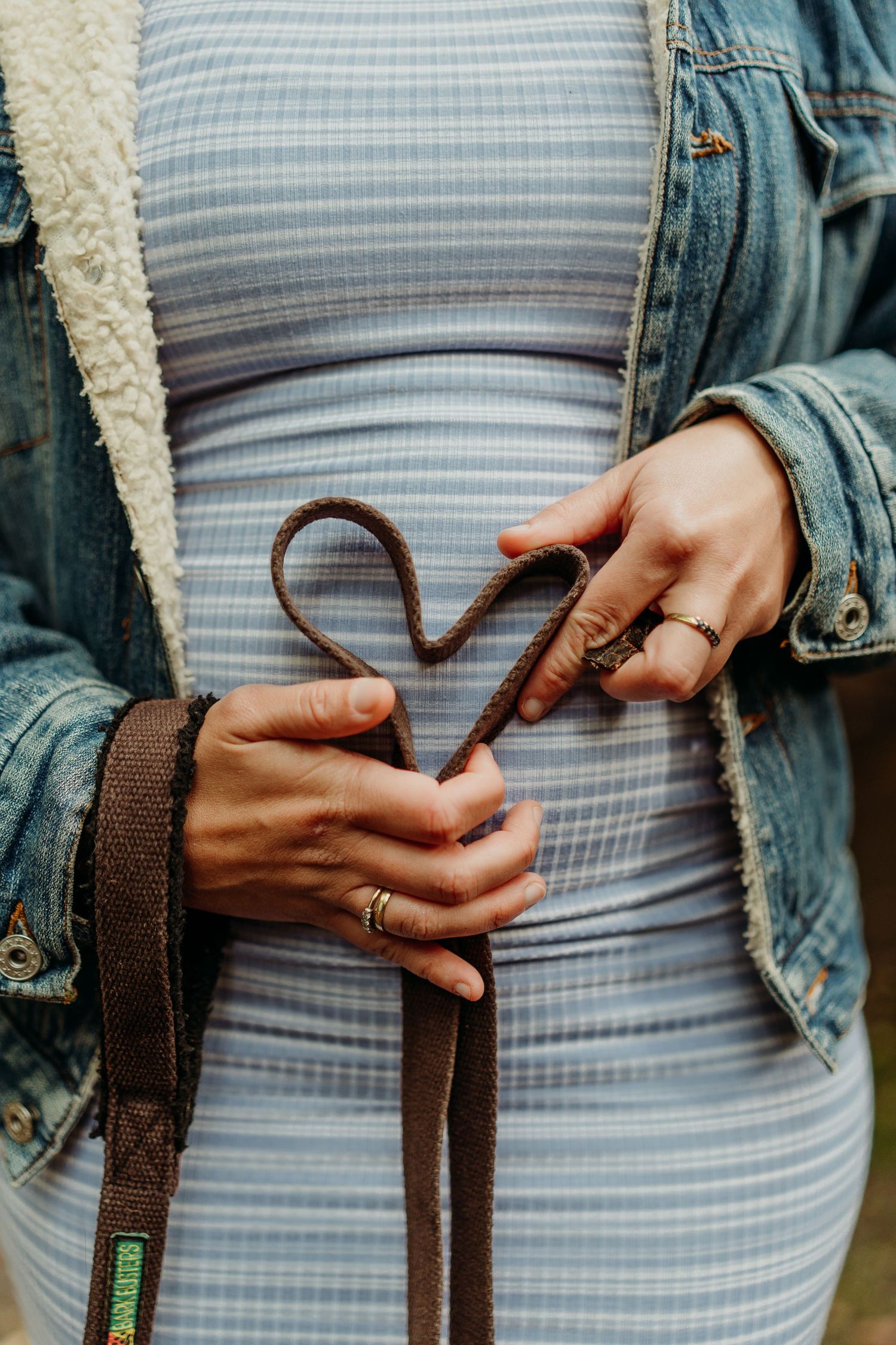 Pregnant woman in striped dress and denim jacket forming a heart shape with a brown leather dog leash.