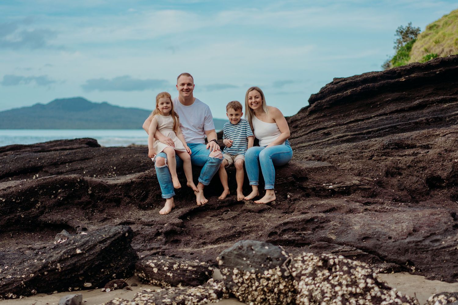 Family sits together on rocky coastal outcrop with ocean and mountains visible in background during sunset.