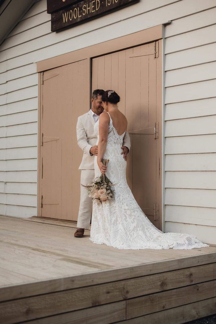 Wedding couple poses against rustic woolshed building with white siding and tan doors.
