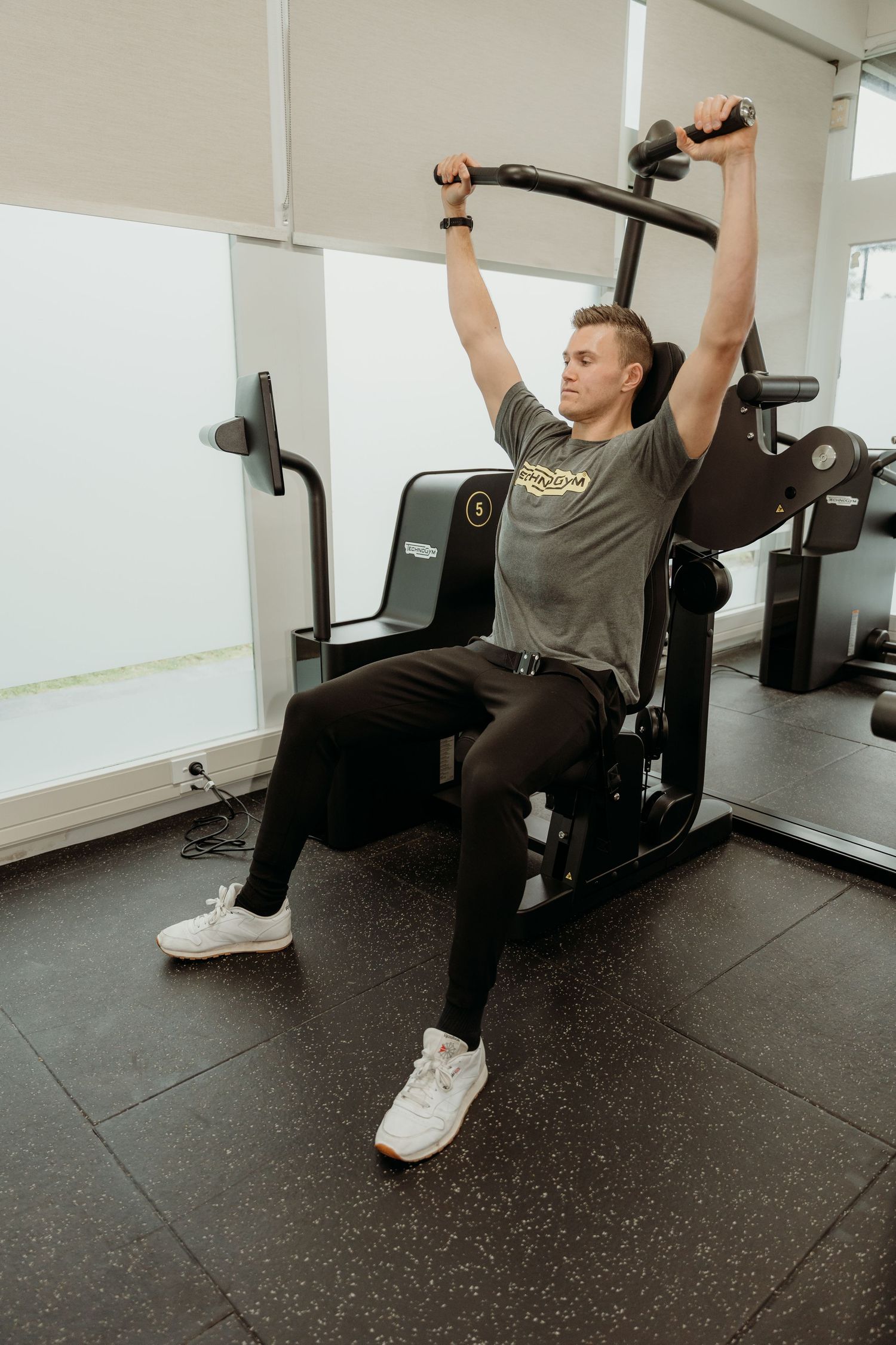 Man performing overhead press exercise on gym machine, wearing gray t-shirt and black pants, in modern fitness center.
