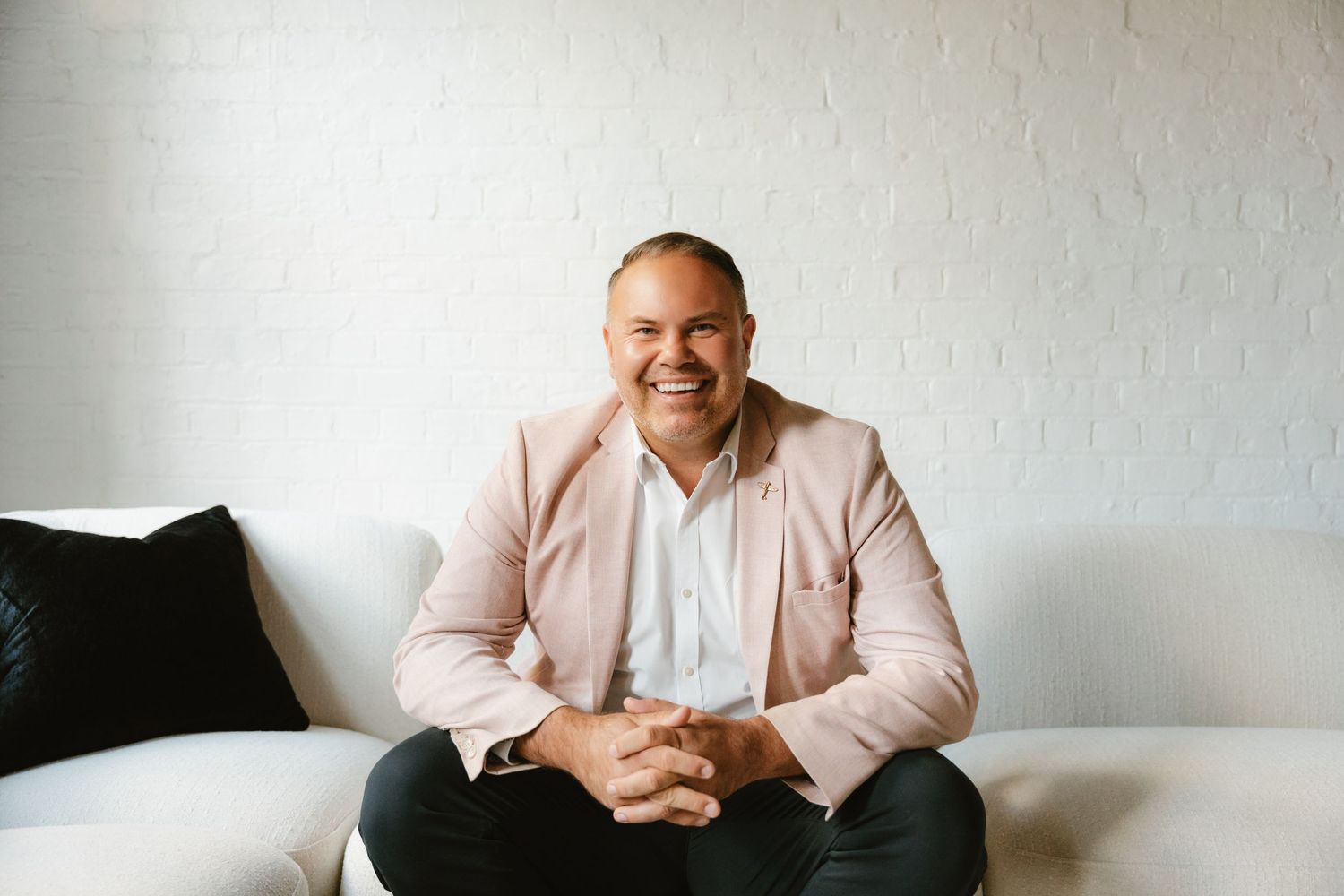 Smiling man in pink blazer sitting cross-legged on white sofa against brick wall.