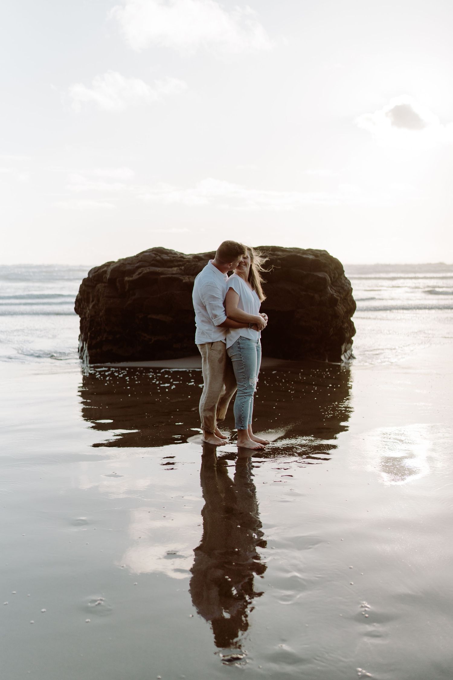 A couple embraces on a misty beach with dark rocks and ocean waves in the background.