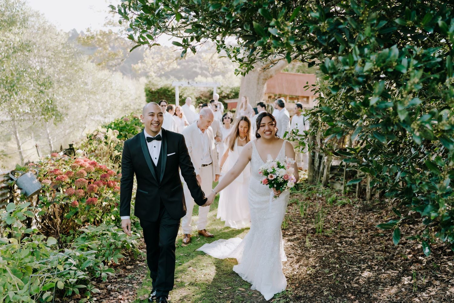 A wedding party walks along a garden path surrounded by lush greenery and flowers.