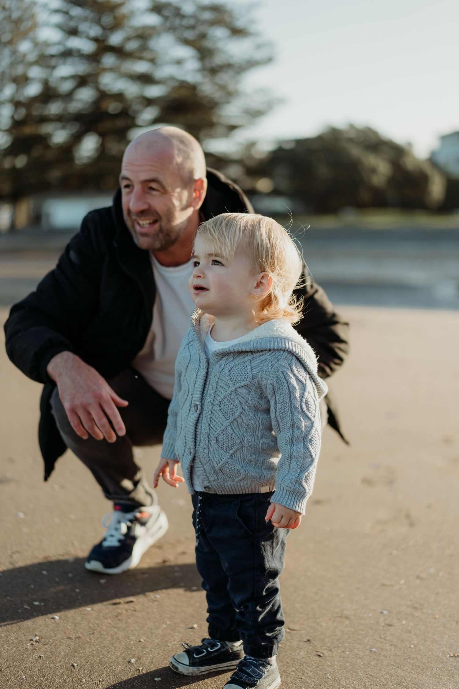 A smiling bald man crouches behind a toddler in a knit sweater on a sandy beach with trees in background.