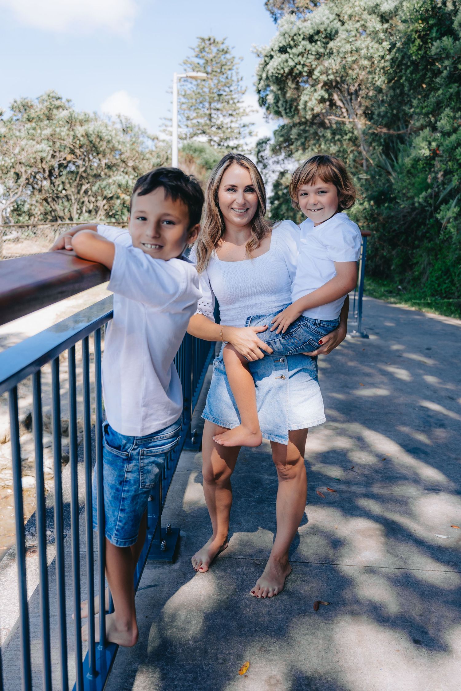 A smiling woman in white top and denim shorts holds a young child while standing beside an older boy on a sunny outdoor path.