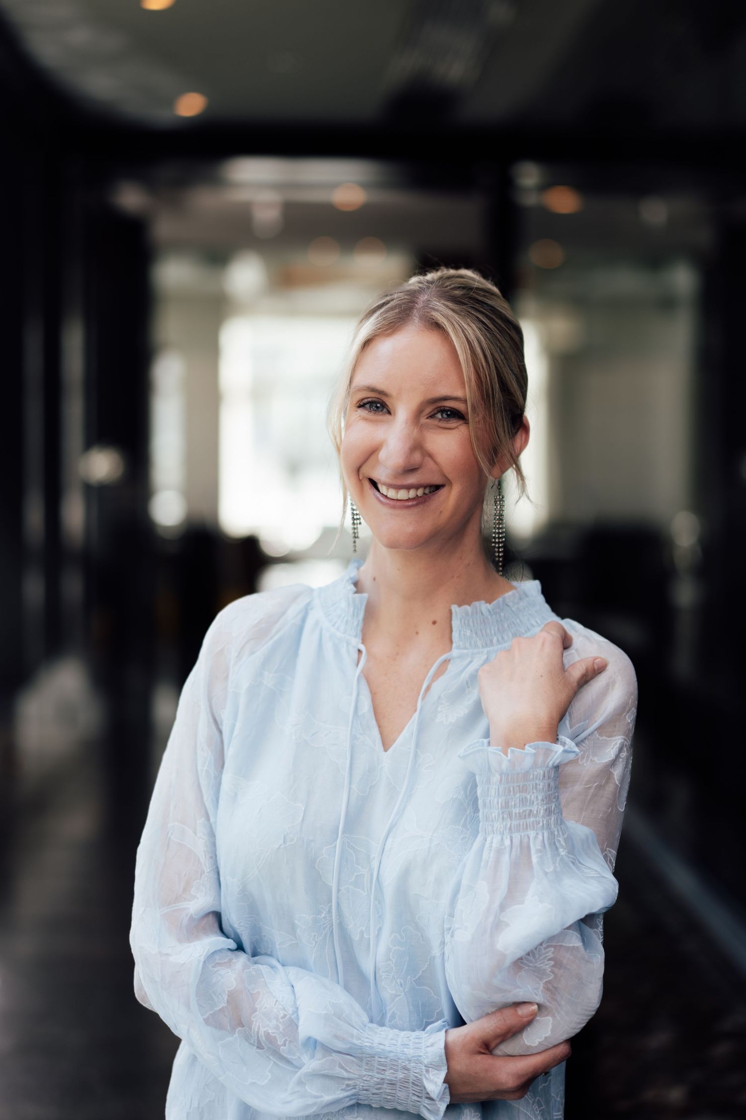 Smiling blonde woman in a light blue long-sleeve blouse with dangling earrings standing in a modern hallway.