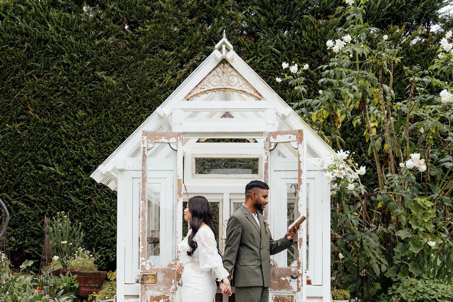 Bride and groom standing before a charming white vintage greenhouse surrounded by lush greenery on their wedding day.