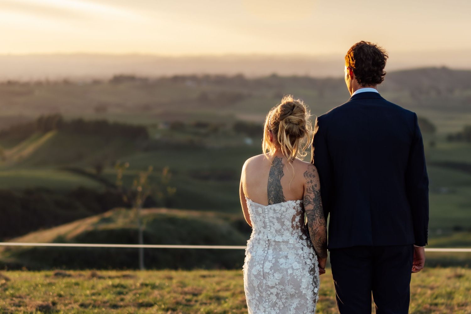 Bride and groom standing together overlooking scenic rolling hills at golden sunset during outdoor wedding ceremony.