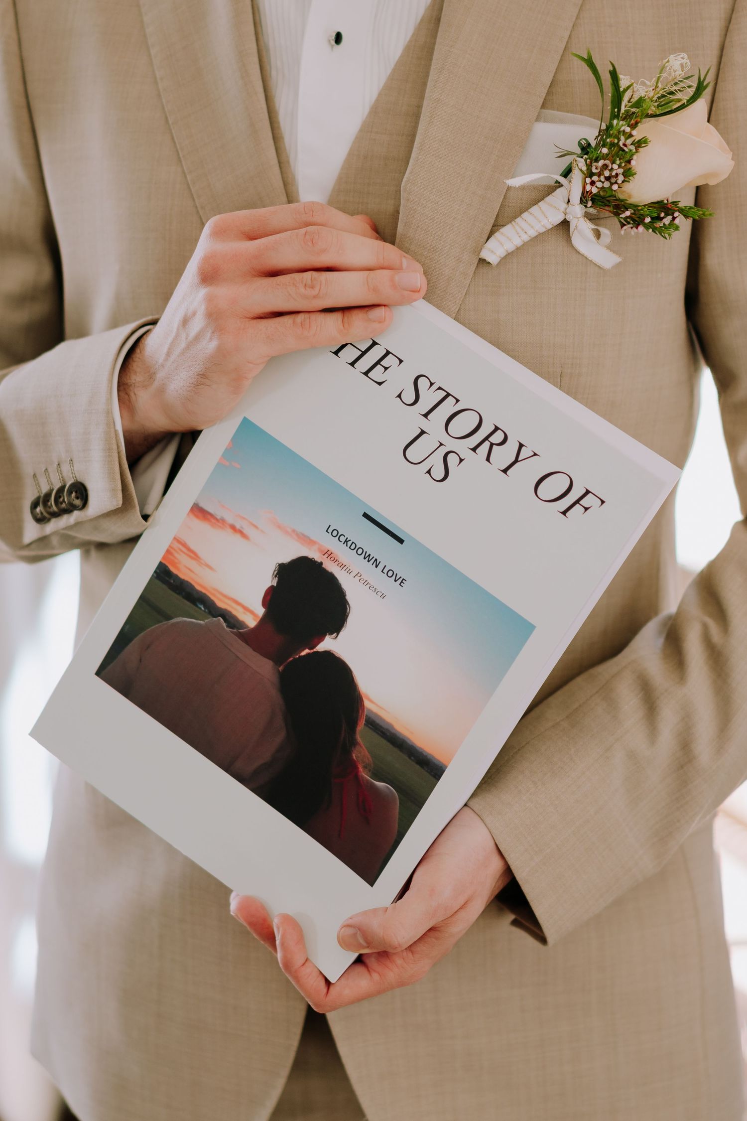 A person in a beige suit holds a wedding photo book titled 'The Story of Us' with a white boutonniere on their lapel.