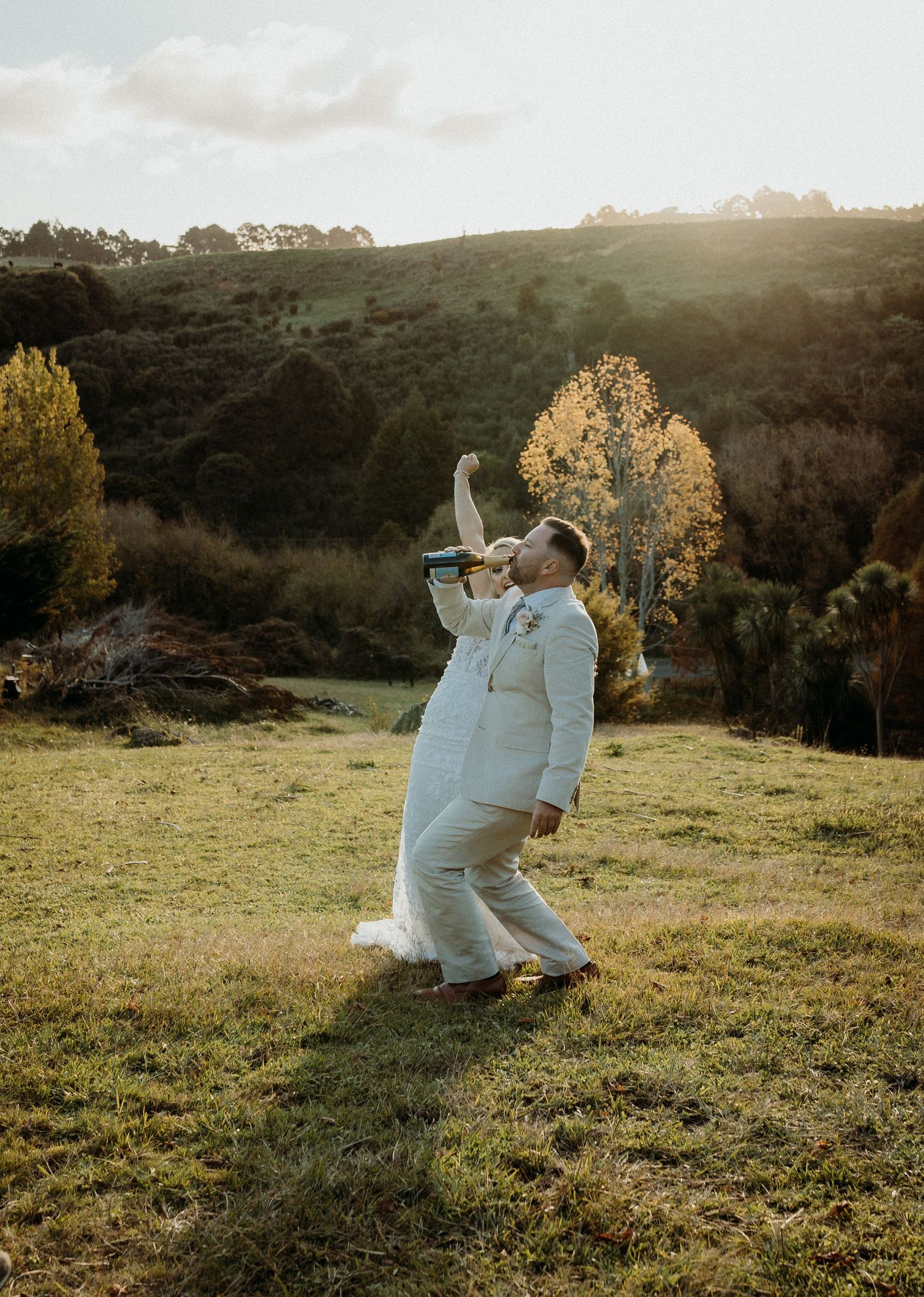 Two people in white outfits sit together on a grassy hillside at sunset enjoying a picnic in nature.