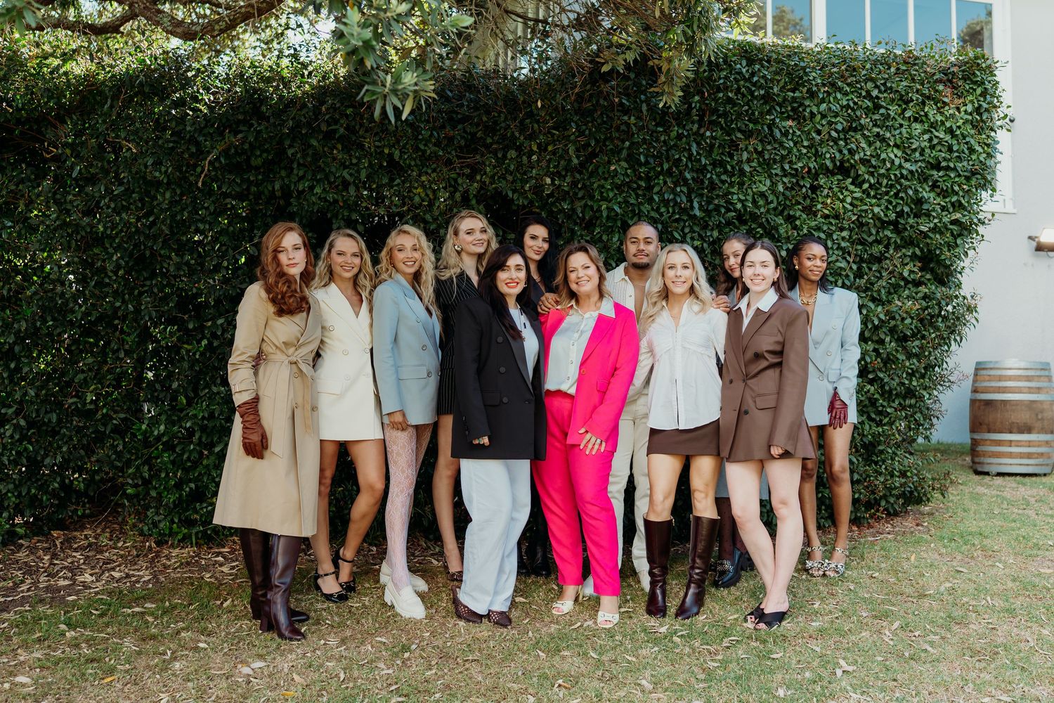 Group of stylish women posing together outdoors in front of a green hedge wall in trendy outfits.