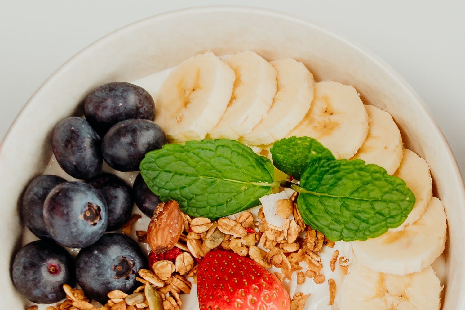 A healthy breakfast bowl topped with blueberries, banana slices, granola, a strawberry, and fresh mint leaves.