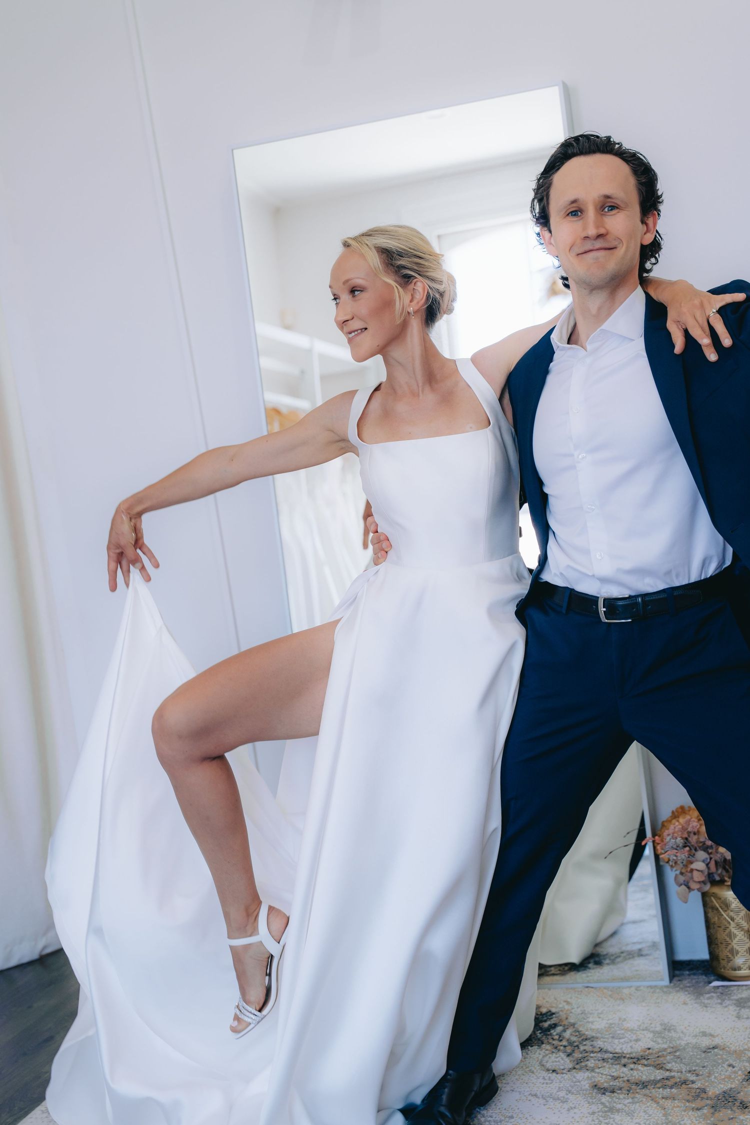 Bride in white gown with slit poses with groom in navy suit near mirror in bright wedding venue.