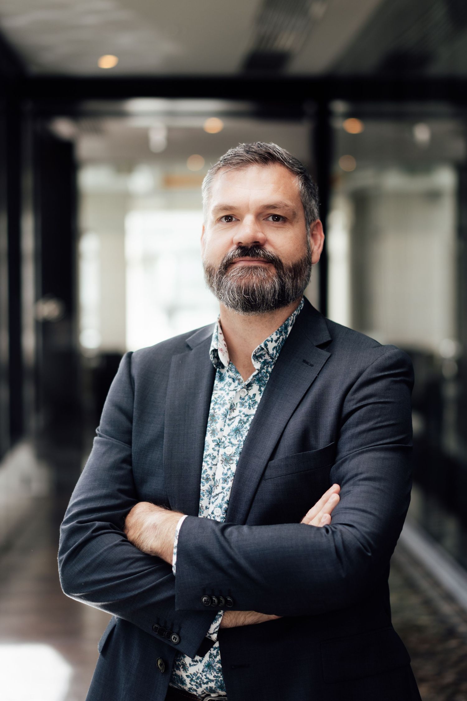 Professional bearded man in dark blazer and floral shirt stands with arms crossed in modern office corridor.