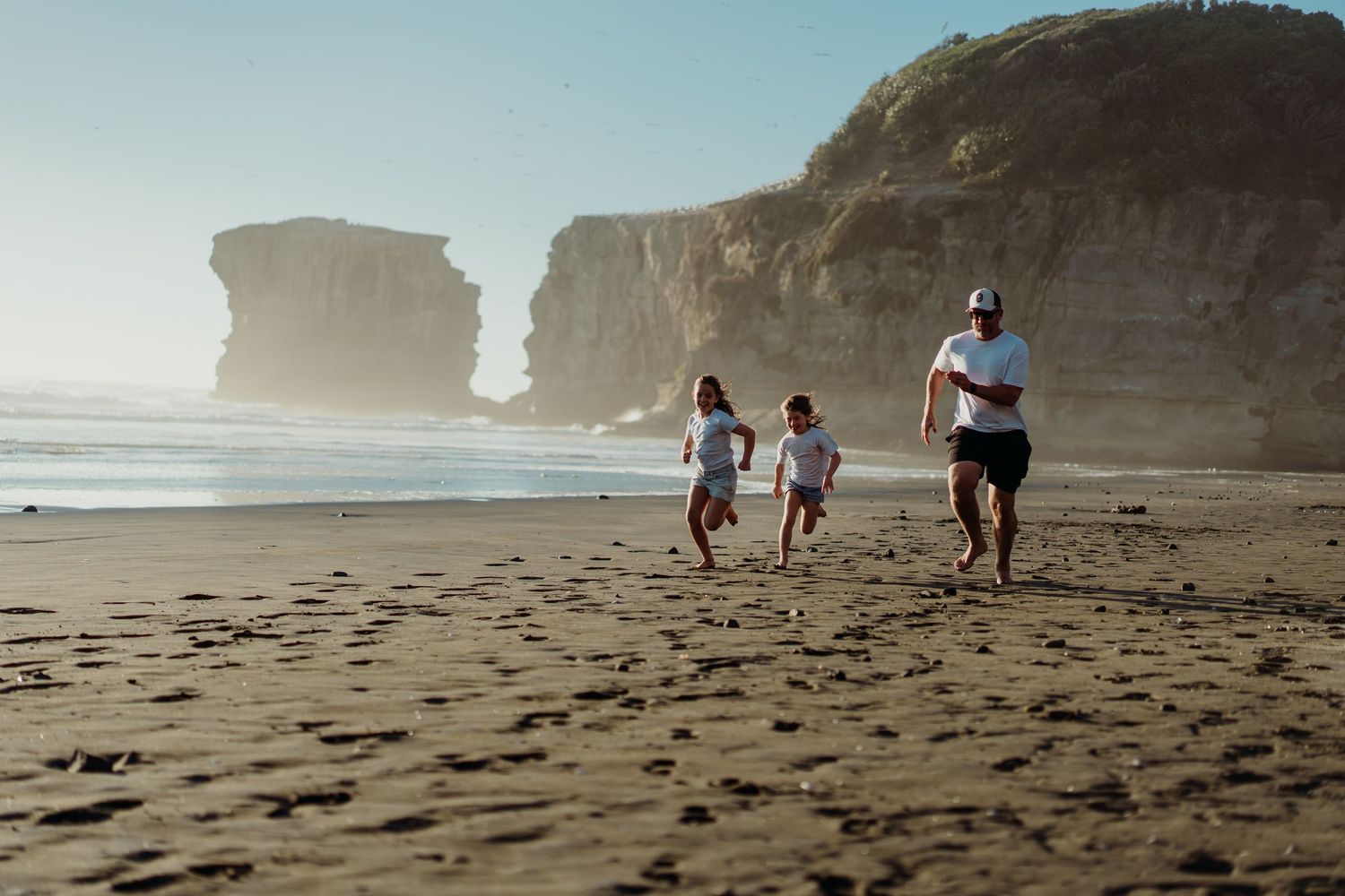 Family running on a sandy beach with dramatic coastal cliffs and ocean waves in the background on a sunny day.