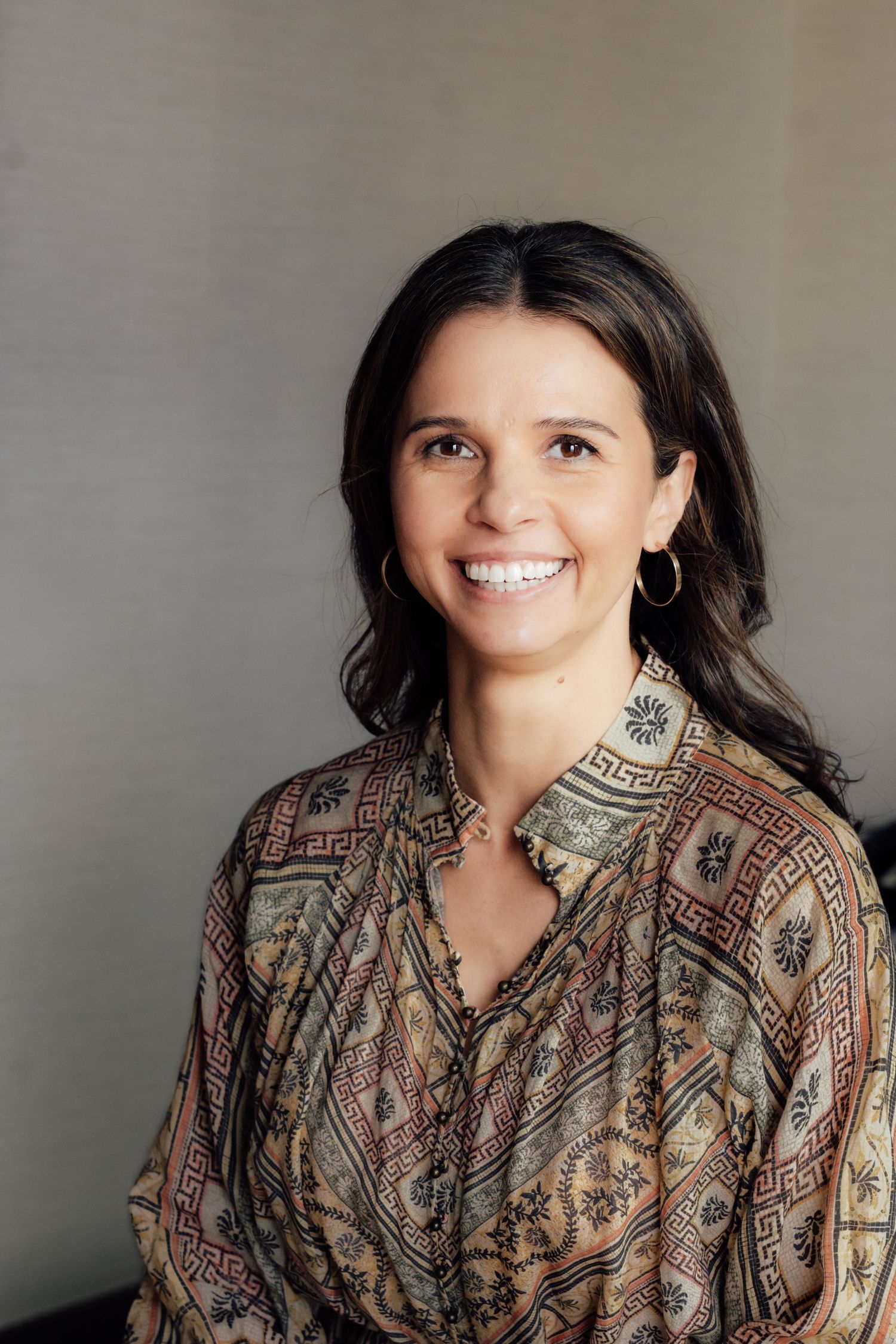Smiling brunette woman wearing gold hoop earrings and a patterned bohemian blouse, posing against a gray background.
