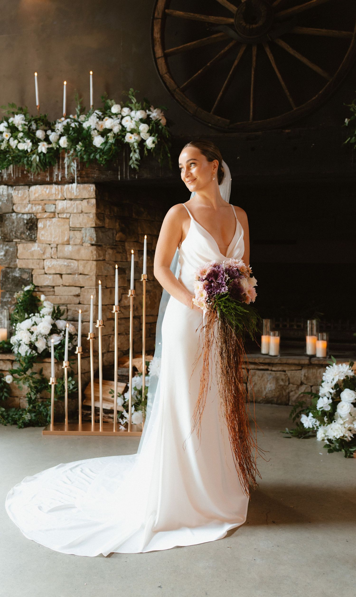 Elegant bridal portrait with white florals and tall candles against rustic stone wall and vintage wagon wheel decor.