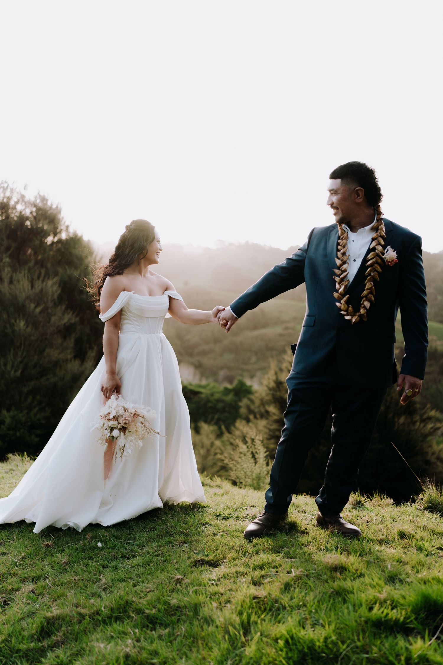 A couple in wedding attire sits on a grassy hilltop during sunset, sharing a playful moment together.