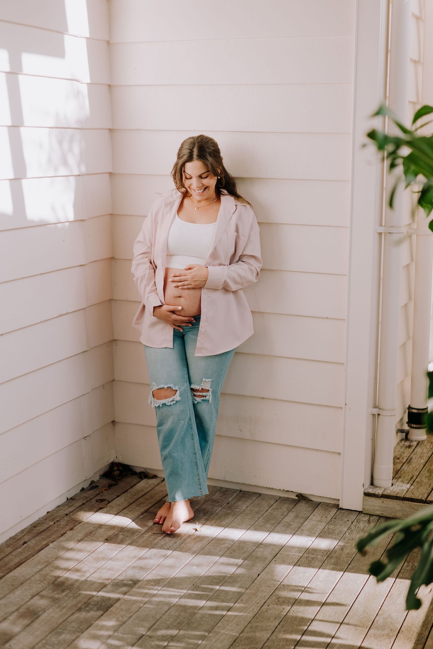 Expecting mother in ripped jeans and pink cardigan stands in sunlit room touching her baby bump.
